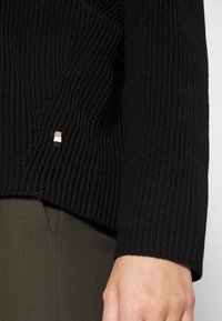 Czarny sweter z dzianiny o prążkowanej fakturze, z małą wielokolorową metką z logo na krawędzi. W połączeniu z ciemnozielonymi spodniami.