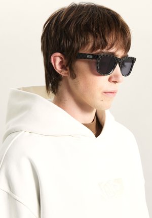 Sorte og grå ternede solbriller med mørke linser, båret sammen med en hvid hættetrøje og en brun skjorte, har et minimalistisk design og klare linjer.