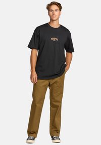 T-shirt negra de algodón con logo naranja, combinada con pantalones beige. Modelo vestido con zapatillas en blanco y negro, de pie frente a un fondo blanco.