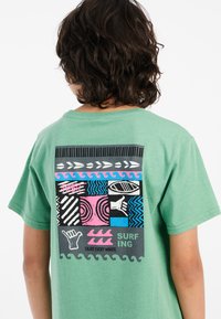 Camiseta de algodón verde con un diseño gráfico colorido en la parte trasera que incluye patrones, olas y el texto: "SURFING" y "ENJOY EVERY MINUTE."