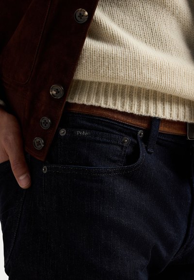 Temno modri jeans iz denima z teksturiranim finišem, delno vidni rjav usnjen pas in krema pulover iz pletenine pod rjavim jopičem.