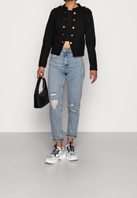 Giacca nera cropped con bottoni dorati, abbinata a jeans strappati blu chiaro e sneakers bianche. Tiene una piccola borsa nera.