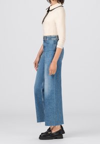 Kvinna som bär en krämfärgad långärmad topp med svart krage och slips, högmidjade vida blåjeans med rå kant, samt svarta loafers med guld detaljer.