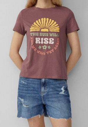 Woman wearing a mauve t-shirt with a yellow sun graphic and text "The sun will rise and we will try again," paired with distressed denim shorts.