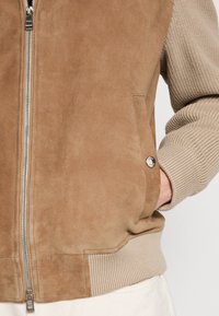 Veste en daim marron clair avec manches et taille en maille côtelée, fermeture éclair argentée et poche avec bouton métallique, main dans la poche.