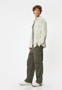 Lys beige skjorte med knapper over en mørkegrøn t-shirt, parret med grønne cargo bukser og hvide sneakers. Funktioner inkluderer store lommer.