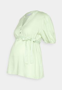 Blouse de maternité vert menthe avec manches bouffantes, fermeture boutonnée et ceinture à nouer à la taille empire. Tissu doux et léger.