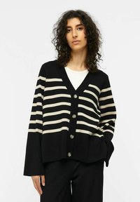 Cardigan a righe nero e crema con scollatura a V, cinque bottoni, maniche larghe e vestibilità rilassata. Il materiale sembra lavorato a maglia con una texture morbida.