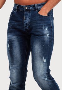 Mörkblå denimjeans med slim fit, som har slitna detaljer, diskret blekning och silverfärgade detaljer vid midjan.