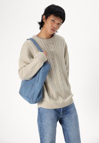 Beigen langhaarigen Pullover mit Rippbündchen, kombiniert mit hellblauen Jeans und einer blauen Canvas-Tasche mit seitlichen Nähdetails.