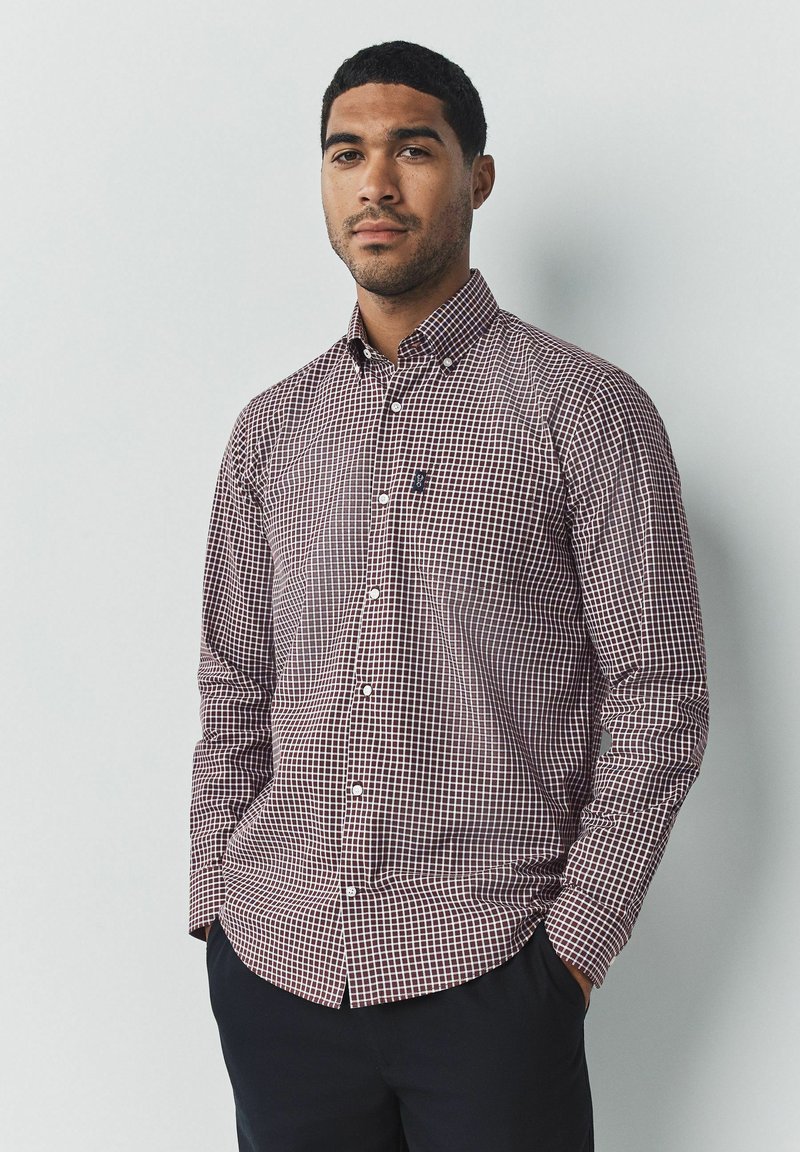 Camisa de botón con un pequeño patrón de cuadros en burdeos y blanco, con mangas largas y un cuello abotonado, combinada con pantalones oscuros.