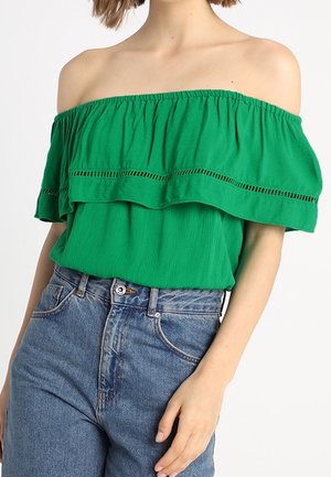 Vrouw met een groene off-shouldertop met ruchesdetails en een blauwe high-waisted spijkerbroek op een witte achtergrond.