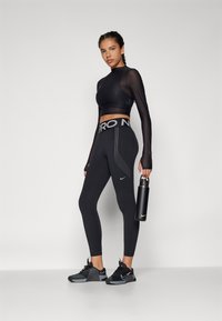 Černý sportovní outfit skládající se z dlouhého sítového crop topu s dlouhým rukávem a legín s vysokým pasem se stříbrnými bočními akcenty. Tenisky a láhev na vodu jsou součástí.