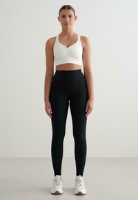 Biele rebrové crop top s výstrihom do V a úzkymi ramienkami, spárovaný s čiernymi legínami s vysokým pásom. Topánky sú biele s sivými akcentmi.