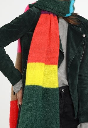 Personne portant une veste en daim vert foncé, un pull gris, un pantalon noir et une écharpe tricotée multicolore avec des rayures rouges, jaunes et vertes.