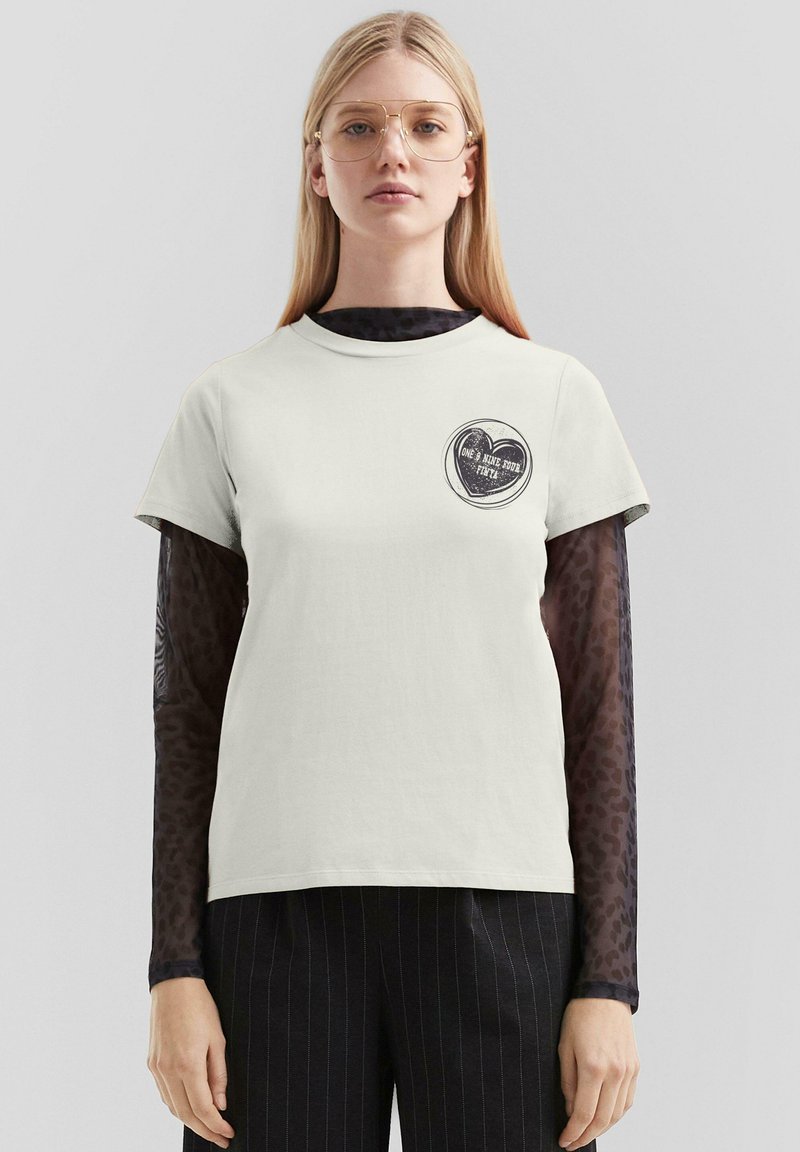 Hellgraues Baumwoll-T-Shirt mit kurzen Ärmeln, mit einem schwarzen runden Grafikdesign auf der linken Brust. Getragen über einem schwarzen gemusterten Langarmoberteil.