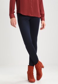 Marinblå snäva byxor i elastiskt tyg, i kombination med rustfärgade mocka ankle boots. Skjortan är mörkröd och har en knapp-up-design.