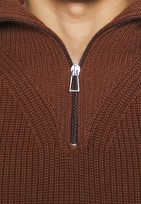 Närbild av en brun stickad tröja med en delvis dragkedjad silverdragkedja vid halsringningen, som visar hud ovanför kragen.