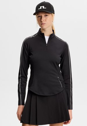 Frau trägt ein schwarzes Langarm-Zip-Up-Oberteil mit weißen Streifen an den Ärmeln, einen schwarzen plissierten Rock und eine schwarze Mütze mit weißem Logo.