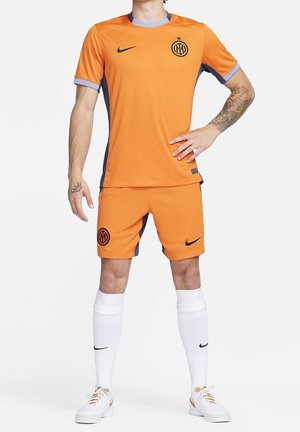 Mand iført orange fodboldtrøje og shorts med sorte logoer, hvide knæstrømper og hvide sneakers, stående med den ene hånd på hoften.