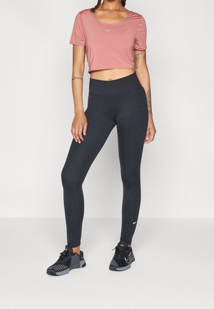 Donna che indossa una maglietta corta Nike rosa, leggings neri a vita alta e scarpe sportive nere, in piedi contro uno sfondo semplice.