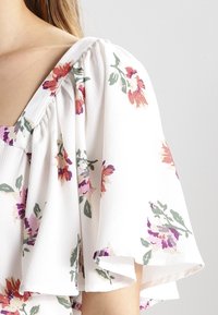 Blusa blanca con patrón floral en tonos de púrpura, rosa y verde. Presenta un escote redondo y mangas cortas con volantes. Tejido texturizado.