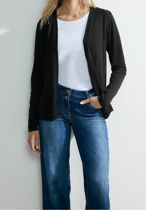 Woman wearing a black open cardigan, white round-neck shirt, and blue high-waist jeans with hand in pocket against plain background.