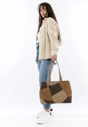 Sac fourre-tout en patchwork marron et beige avec une texture lisse, des poignées doubles et un pendentif, associé à un jean clair et des baskets blanches.