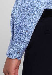 Jasnoniebieski rękaw koszuli z białym kwiatowym wzorem, z mankietami zapinanymi na guziki i gładką tkaniną, zestawiony z czarnymi spodniami.