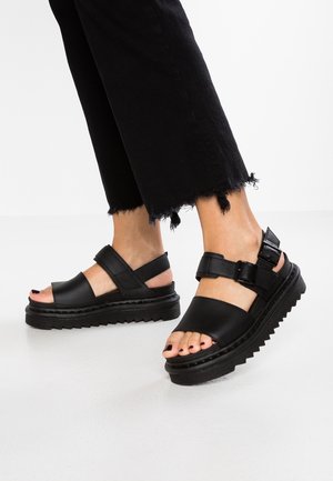 Feet wearing black platform sandals with thick soles and ankle straps, paired with black cropped pants with frayed hems.