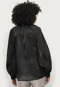 Blusa in raso nero con maniche lunghe e voluminose. Presenta un collo alto e dettagli arricciati sul retro, pannello inferiore trasparente.