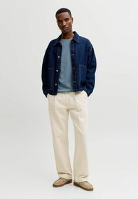 Homme portant une veste en jean bleue, une chemise bleu clair, un pantalon crème et des chaussures beiges, debout les mains dans les poches sur fond blanc.