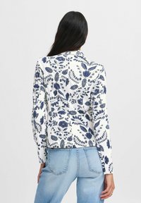 Giacca a stampa floreale bianca con fiori e foglie blu navy. Maniche lunghe, design leggermente cropped, texture liscia e collo a camicia.
