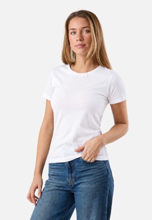 Jeune femme aux longs cheveux blonds portant un t-shirt blanc uni à manches courtes et un jean bleu sur un fond neutre.