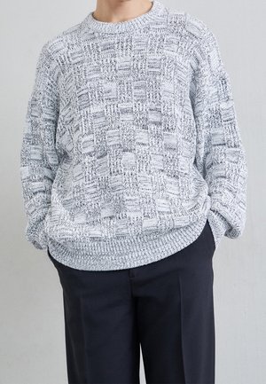 Hellgrauer, gestrickter Pullover mit einem strukturierten, gitterartigen Muster und geripptem Saum. Getragen über dunklen Hosen mit einem Rundhalsausschnitt.