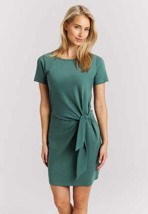 Vrouw met blond haar die een groene jurk met korte mouwen draagt, gestrikt in een knoop ter hoogte van de taille, staand tegen een eenvoudige lichte achtergrond.