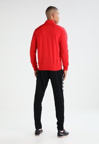 Rode sportjasje met een hoge kraag en lange mouwen, gecombineerd met zwarte broek met witte chevronaccenten. Zachte textuur.