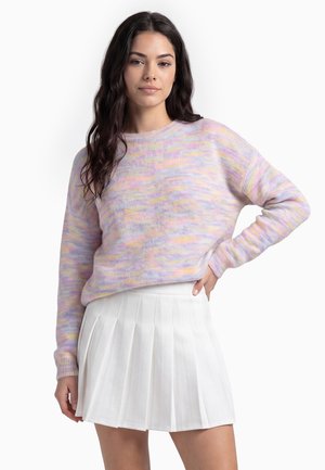 Kvinde med langt mørkt hår iført en pastel flerfarvet sweater og et hvidt plisseret skørt, stående med en hånd på hoften.