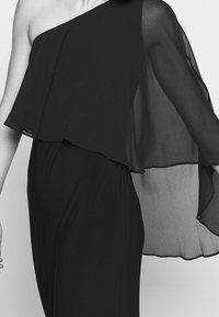 Svart enaxlad topp med lätt, genomskinligt tyg och draperad design. Det flödande materialet skapar en lagerpåläggseffekt.