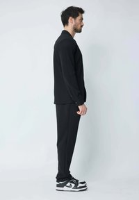Chemise noire à manches longues avec des boutons aux poignets, associée à un pantalon noir assorti et des baskets noir et blanc, vue de profil.