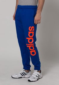 Modré tepláky s širokým oranžovým logem "adidas" na levé noze. Vyrobené z měkké texturované tkaniny, s elastickým pasem.