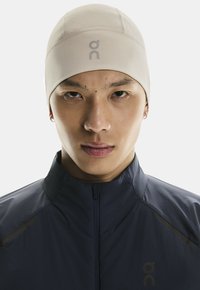 Casquette de course en tissu stretch beige au design épuré, arborant un petit logo et une forme ajustée, portée avec une veste zippée bleu marine.