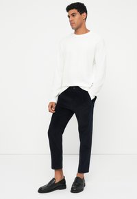 Biela košeľa s dlhým rukávom, námornícke kordové nohavice a čierne kožené slip-on topánky. Hladké textúry a priliehavá silueta.