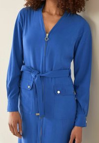 Blauwe jurk met een rits aan de voorkant, lange mouwen, verstelbare tailleband en twee borstzakken met zilveren knopen. Gladde stofstructuur.