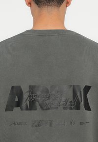 Šedá mikina s výrazným černým logo "ARKK" a menším textem pod ním. Tkanina má strukturovaný vzhled s žebrovaným výstřihem.