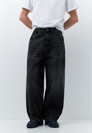 Osoba v čiernych voľných džínsoch, čiernych topánkach a obyčajnom bielom tričku stojaca pred jednoliatym svetlým pozadím.