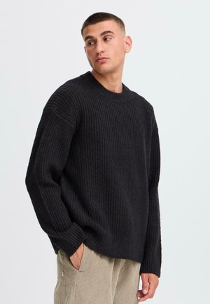 Schwarzer gerippter Strickpullover mit rundem Halsausschnitt und überschnittenen Schultern. Der Stoff wirkt strukturiert und gemütlich. Das Model posiert mit den Händen in den Taschen.