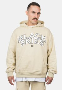 Beige mikina s výrazným čiernym nápisom "ČIERNE NEBESÁ", klokaní vrecko, ribované manžety a voľný strih. Hladký bavlnený materiál.