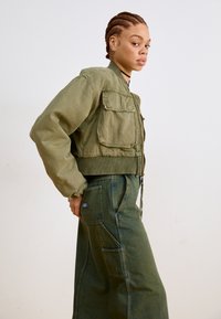 Oliivinvihreä cropped bomber-takki, jossa on ribbetyt hihansuut ja kaksi etutaskua, yhdistettynä korkeavyötäröisiin tummanvihreisiin leveälahkeisiin farkkuihin.