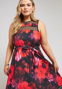 Vestido con estampado floral que presenta un fondo negro, flores rojas y rosas, un yugo transparente y una cintura ceñida con un lazo de cinta.